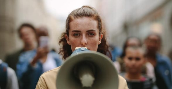 A fair-skinned, dark-haired person holds a megaphone to their mouth. They're joined by out-of-focus protestors in the background.