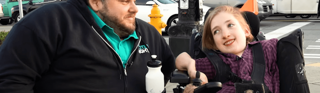 man in black sweatshirt and woman with purple jacket sitting in wheelchair talking on a sidewalk