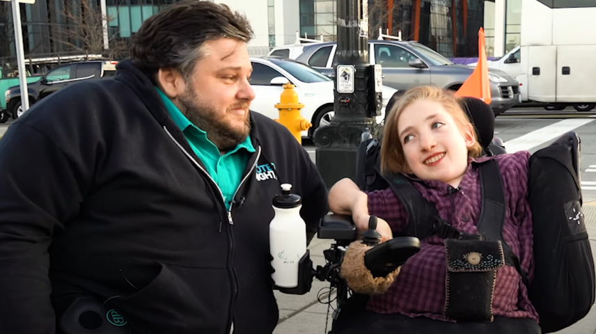man in black sweatshirt and woman with purple jacket sitting in wheelchair talking on a sidewalk