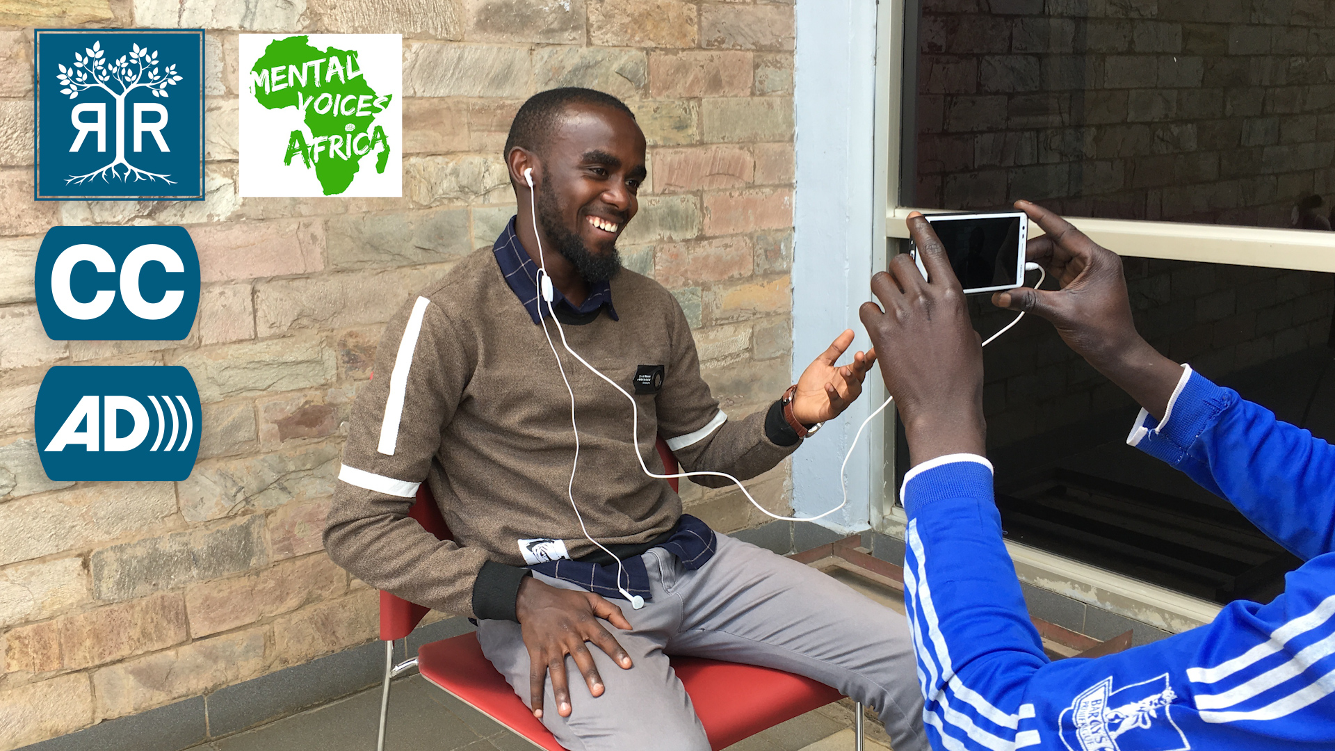 A photo of Brian seated in a chair with headphones in his ears looking toward a mobile phone being held toward him.