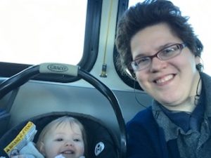 Ivanova is smiling into the camera. To the left is a smiling baby in a car seat.