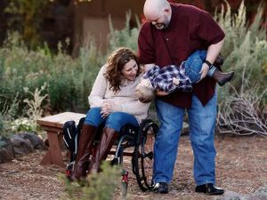 Family photo of Jessica and her family. There are 2 adults and 1 child. They are outside in front of a garden setting and are holding the young child in the photo.