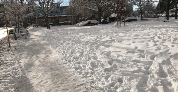 A blanket of snow covers an open space; a sidewalk buried underneath a sheet of icy packed snow.