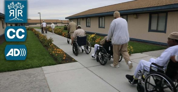 A row of inmates move down a walkway. Several are in wheelchairs. Another uses a walker.