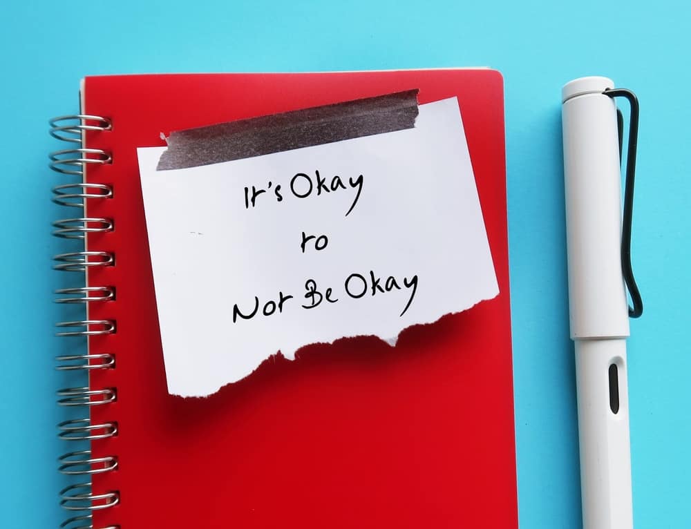 A red, spiral notebook and white pen against a light blue background. A white, torn off post-it note is taped to the notebook. It reads: "It's okay to not be okay."