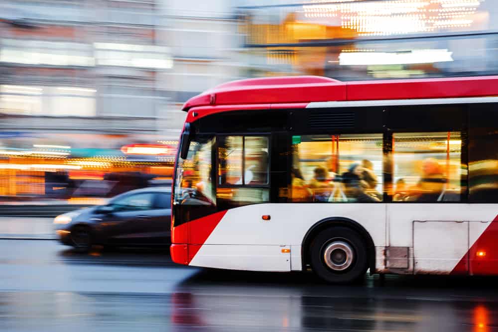 Outdoors, day. A red and white bus in motion in traffic. The buildings and other vehicles around it are a blur.