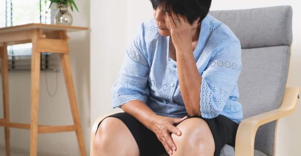 An older Asian woman with short dark hair waits in an office setting. She holds her head in her left hand, looking despondent. She's wearing a short-sleeved light blue top and dark shorts.