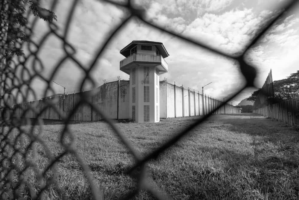 Outdoors, day. A prison watchtower separated by a wired fence. The camera is close up on the fence.