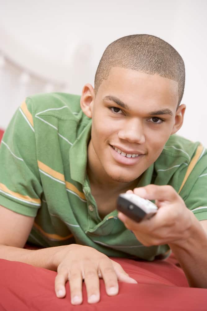 A light-skinned Black teenager in a green, striped, short-sleeved shirt is lying down on a bed, facing the camera and smiling. He has a TV remote in his hand and is pointing it at the camera.