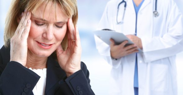 A patient, a blonde white woman wearing a dark top with a white top underneath, holds her head in pain while a fair-skinned doctor, face unseen, holds a clipboard while wearing a white lab coat with a stethoscope around their neck.