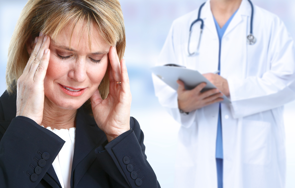 A patient, a blonde white woman wearing a dark top with a white top underneath, holds her head in pain while a fair-skinned doctor, face unseen, holds a clipboard while wearing a white lab coat with a stethoscope around their neck.