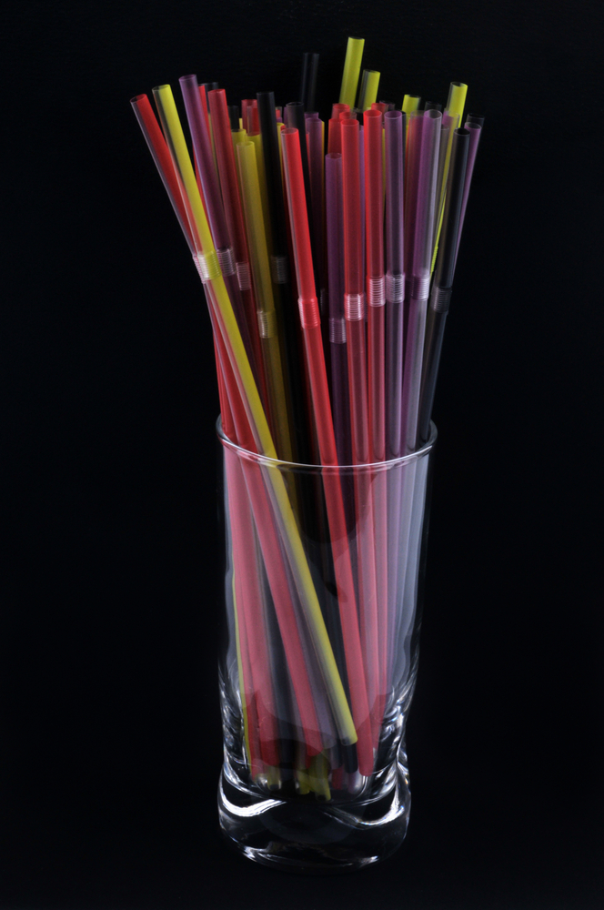 Black background. A clear drinking glass filled with multicolored plastic straws.