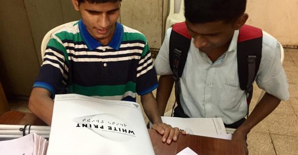 Indoors. Two dark brown-skinned Indian teens, one wearing a bookbag on their back, look down at copies of White Print mag that are located on a brown table.