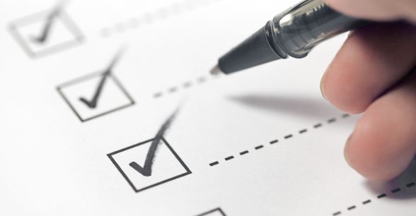 A white piece of paper with several boxes checked off by black check marks. A hand holds a pen above the check marks.