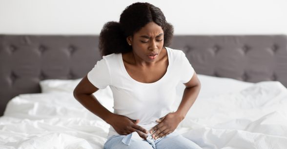 A young, brown-skinned Black woman seated on a bed holds her abdomen in pain.