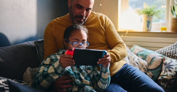 Indoors, day. A father, a light-skinned Black man, and his child, light brown-skinned, sit on a couch together as the child, wearing glasses, plays on an electronic device. The father watches.