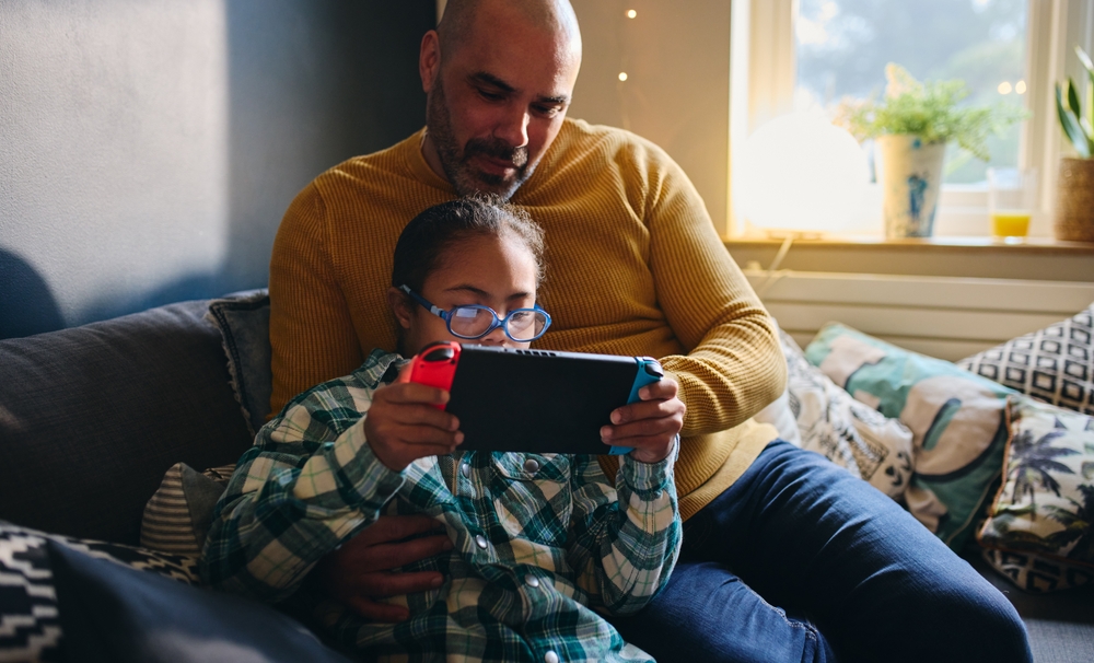 Indoors, day. A father, a light-skinned Black man, and his child, light brown-skinned, sit on a couch together as the child, wearing glasses, plays on an electronic device. The father watches.