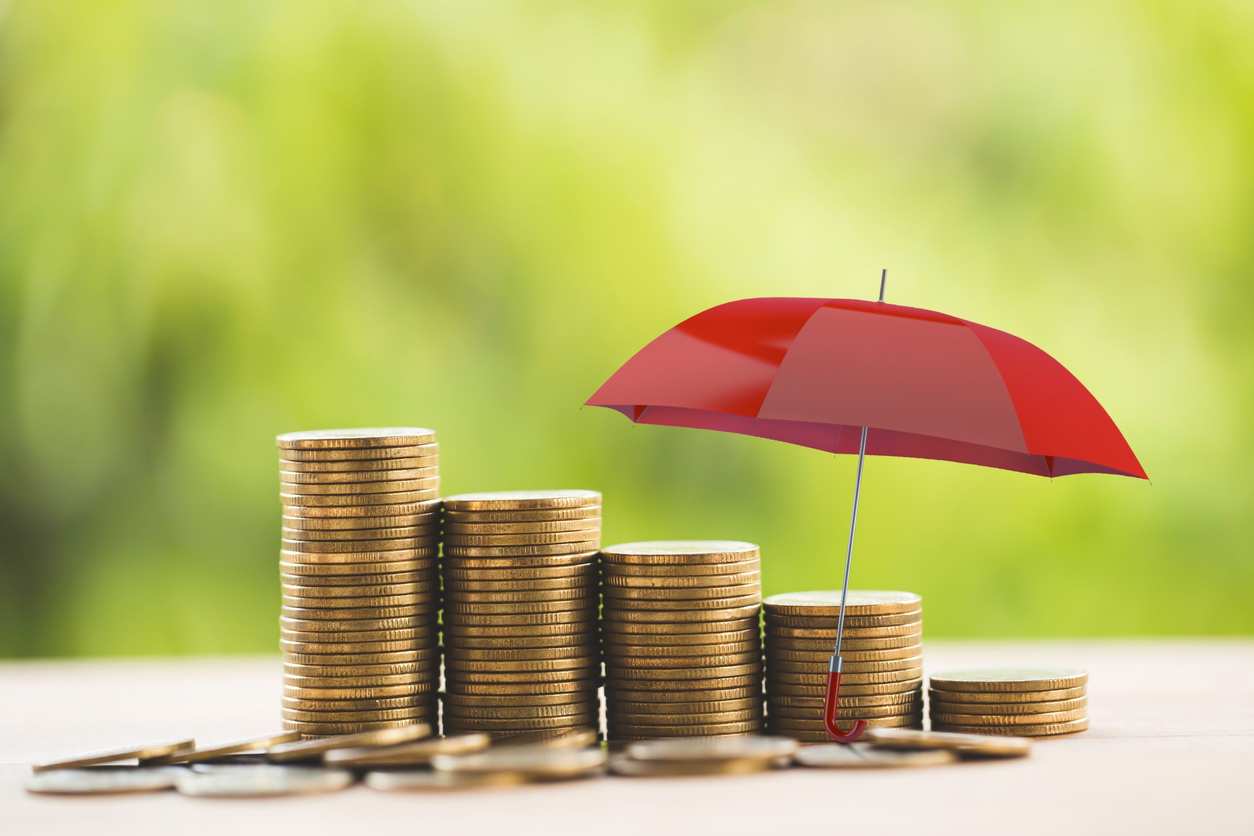 Outdoors, day. A tiny red umbrellla sits on a table. It is partially protecting five stacks of gold coins, organized from left to right from the biggest to the smallest stack. Some unstacked gold coins are strewn around the stacks.