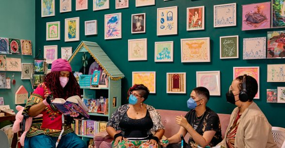 Four masked people of color discuss books in an art-filled bookstore. On the left, a Black non-binary woman has everyone’s attention, pointing out a passage from the book they’re holding while sitting in a rollator with their cane between their legs. Next to them, an Indigenous woman and Latinx non-binary person share a couch. On the right, a Black non-binary pereson sits in a folding chair while holding their own book and fidget stim.