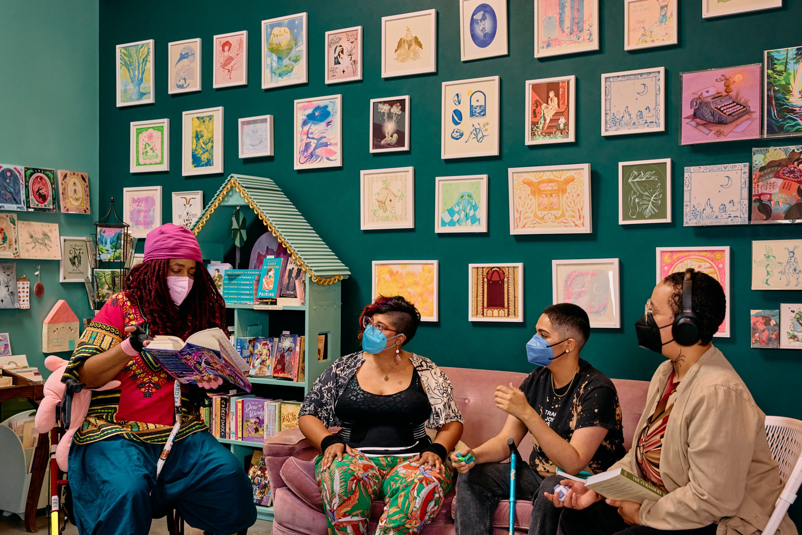 Four masked people of color discuss books in an art-filled bookstore. On the left, a Black non-binary woman has everyone’s attention, pointing out a passage from the book they’re holding while sitting in a rollator with their cane between their legs. Next to them, an Indigenous woman and Latinx non-binary person share a couch. On the right, a Black non-binary pereson sits in a folding chair while holding their own book and fidget stim.