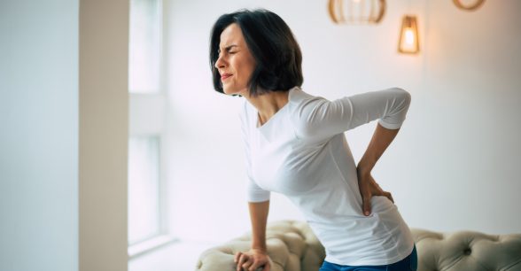 Indoors, day. A white woman with shoulder-length dark hair wearing a white top and jeans is holding her lower back with her right hand, in excruciating pain, while standing in front of a beige couch tightly holding onto its arm with her left hand.