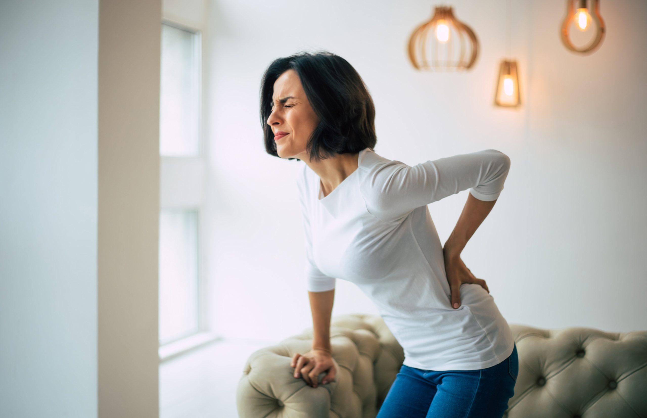 Indoors, day. A white woman with shoulder-length dark hair wearing a white top and jeans is holding her lower back with her right hand, in excruciating pain, while standing in front of a beige couch tightly holding onto its arm with her left hand.