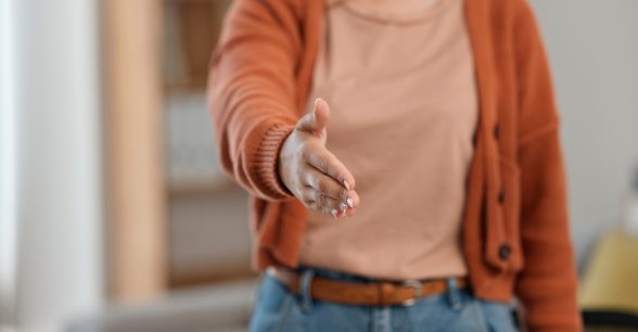 A fair- or possibly light-skinned person seen from the neck to the hips. She's wearing a tan or light pink top with a burnt orange cardigan over it and blue jeans with a light brown belt. With shoulder length dark hair, she reaches out her hand for a handshake and friendly 'Hello."