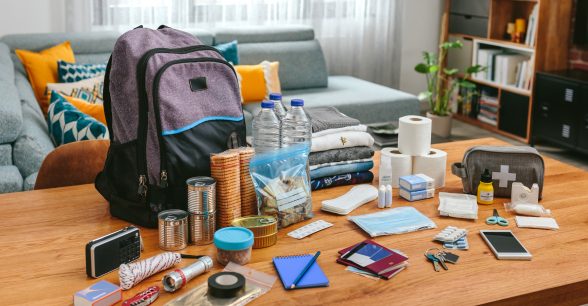 Indoors, day. In a living room, a coffee table sits with a backpack, plastic water bottles, rolls of tissue, a flashlight, crackers, and other assorted items on top of it.
