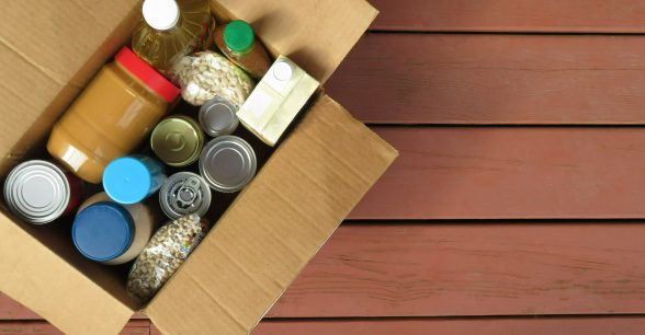 A photo of a cardboard box filled with shelf-stable goods including peanut butter, dry beans, and vegetable oil. The box is placed on some kind of wood paneling like a picnic table or bench.