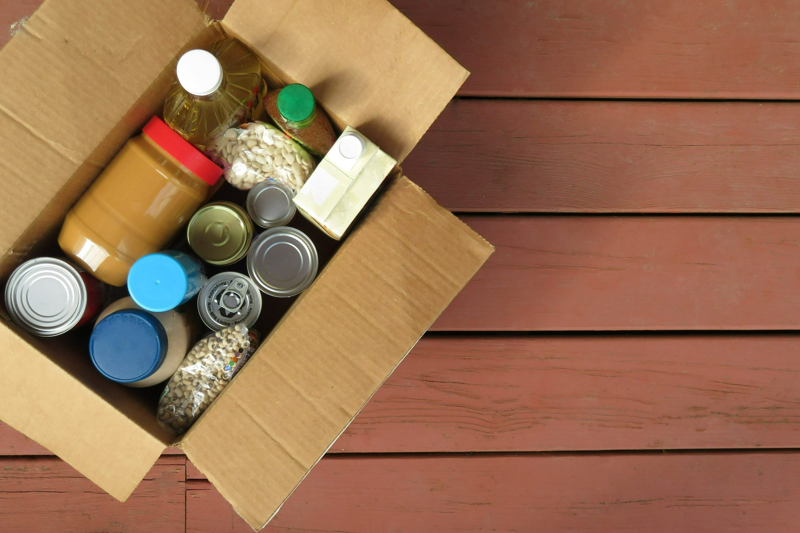 A photo of a cardboard box filled with shelf-stable goods including peanut butter, dry beans, and vegetable oil. The box is placed on some kind of wood paneling like a picnic table or bench.