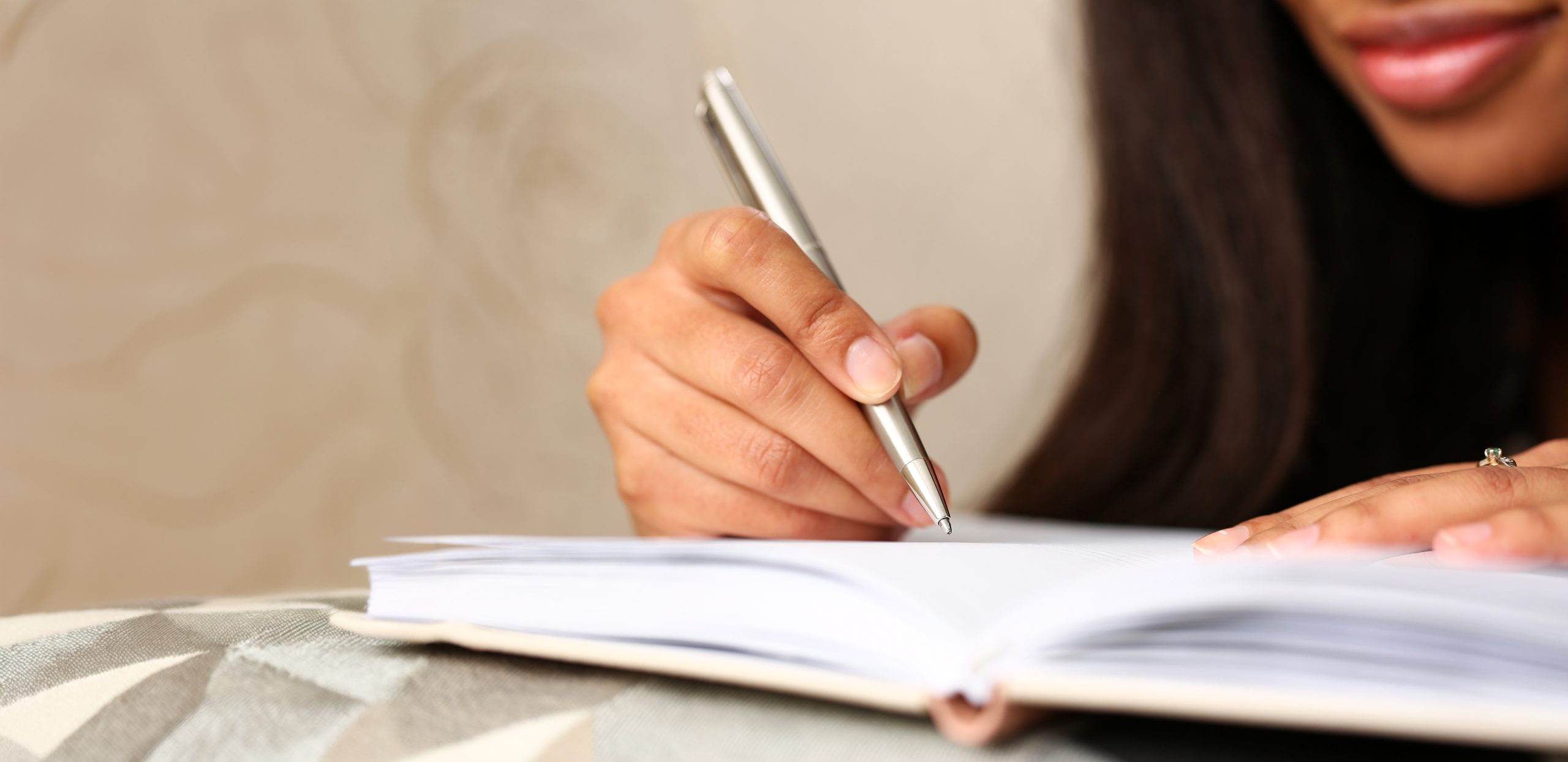 A light brown-skinned Black person with long, straight dark hair has a journal of some kind open in front of them as they use a silver pen to write inside of it.