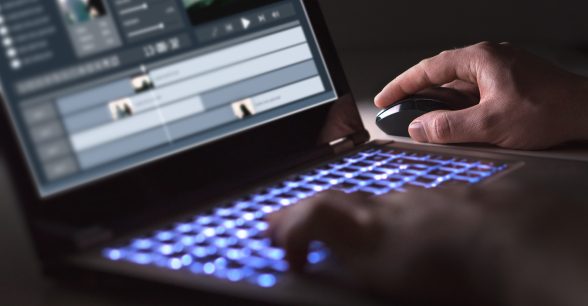 Indoors. Fair-skinned hands of a person are visible at the keyboard of a laptop in a dimly lit room. The keyboard lights up a light blue. The person is in the middle of editing a video with professional editing software. Their right hand holds a black computer mouse while their left hand types on the laptop.