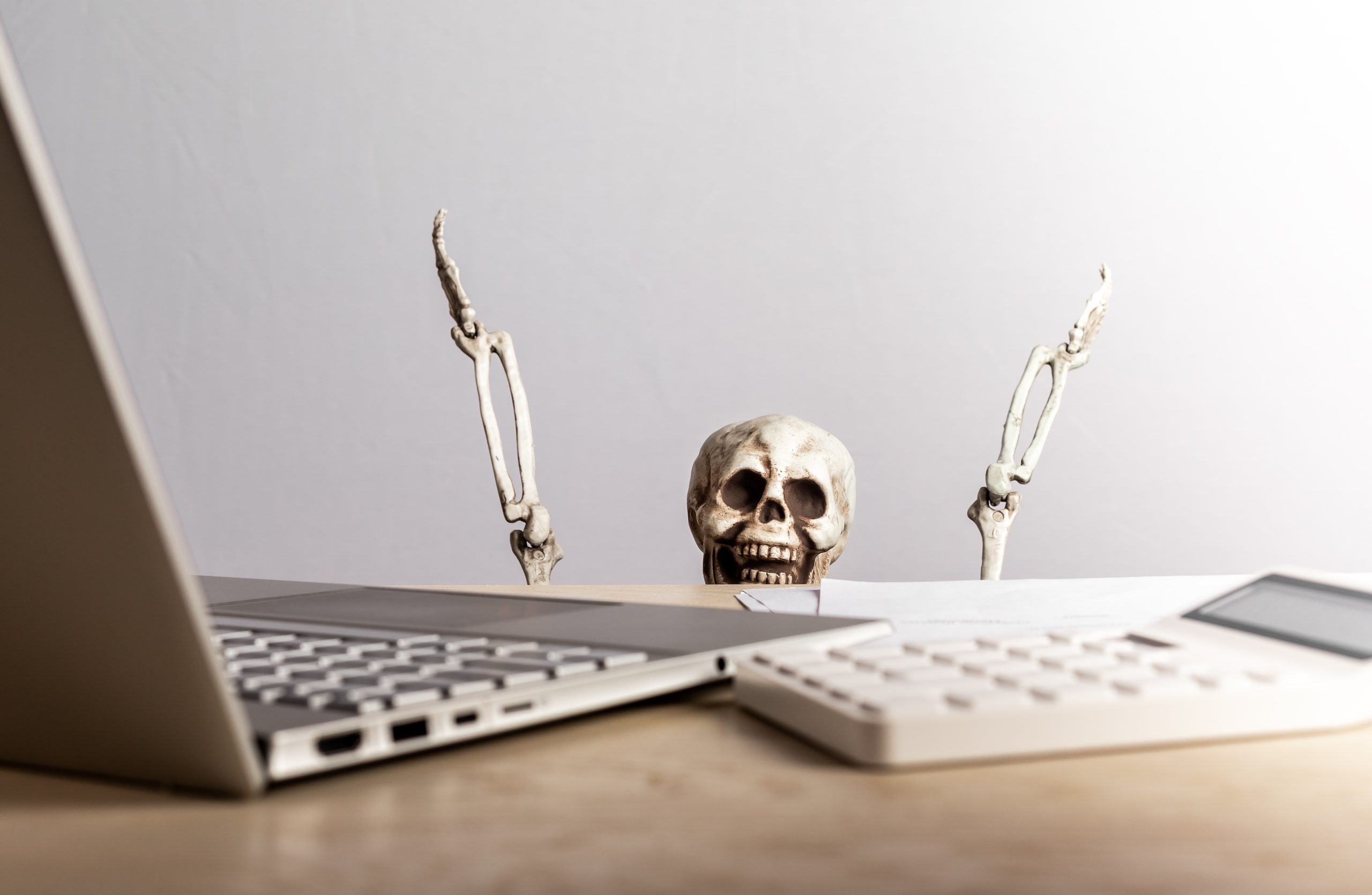 A skeleton behind a desk with its bone arms in the air looking at a laptop, calculator, and pile of papers.
