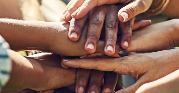 Outdoors. A small group of people with varying skin tones have their hands in a pile together in unity. Only hands, arms, and torsos are visible. Those with visible torsos are dressed casually.