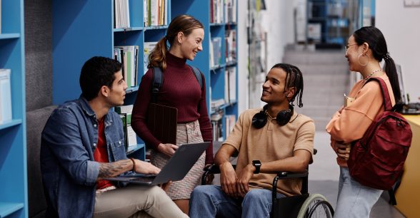 A group of young adult students with various skin colors, phenotypes, and clothing and hairstyles chats cheerfully in a college library, One of the students, a brown-skinned person with short braids, is a wheelchair user.