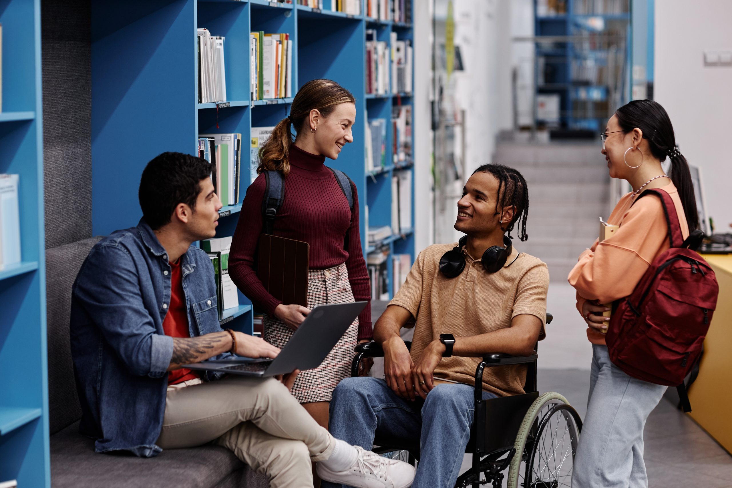 A group of young adult students with various skin colors, phenotypes, and clothing and hairstyles chats cheerfully in a college library, One of the students, a brown-skinned person with short braids, is a wheelchair user.