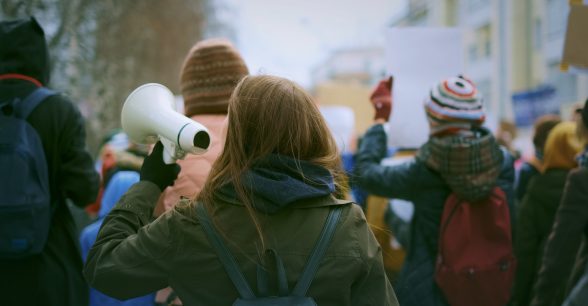 Outdoors, day. A crowd of protestors seen from behind. Some people are wearing winter hats and backpacks. A person with long brown hair in the center holds up a blow horn.
