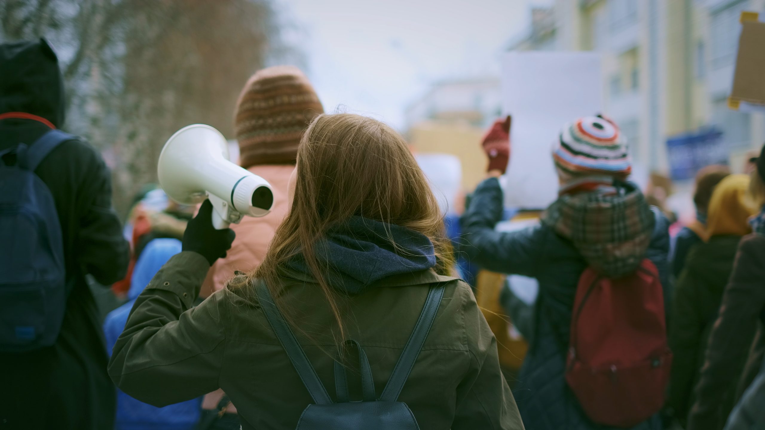 Outdoors, day. A crowd of protestors seen from behind. Some people are wearing winter hats and backpacks. A person with long brown hair in the center holds up a blow horn.