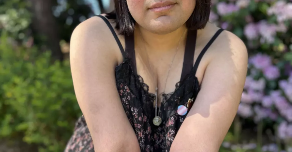 A photo of Angela outdoors leaning forward with arms crossed. She wear a trans pride pin. In the background, sunlight highlights green grass, light pink flowers, and tall trees against a blue sky.