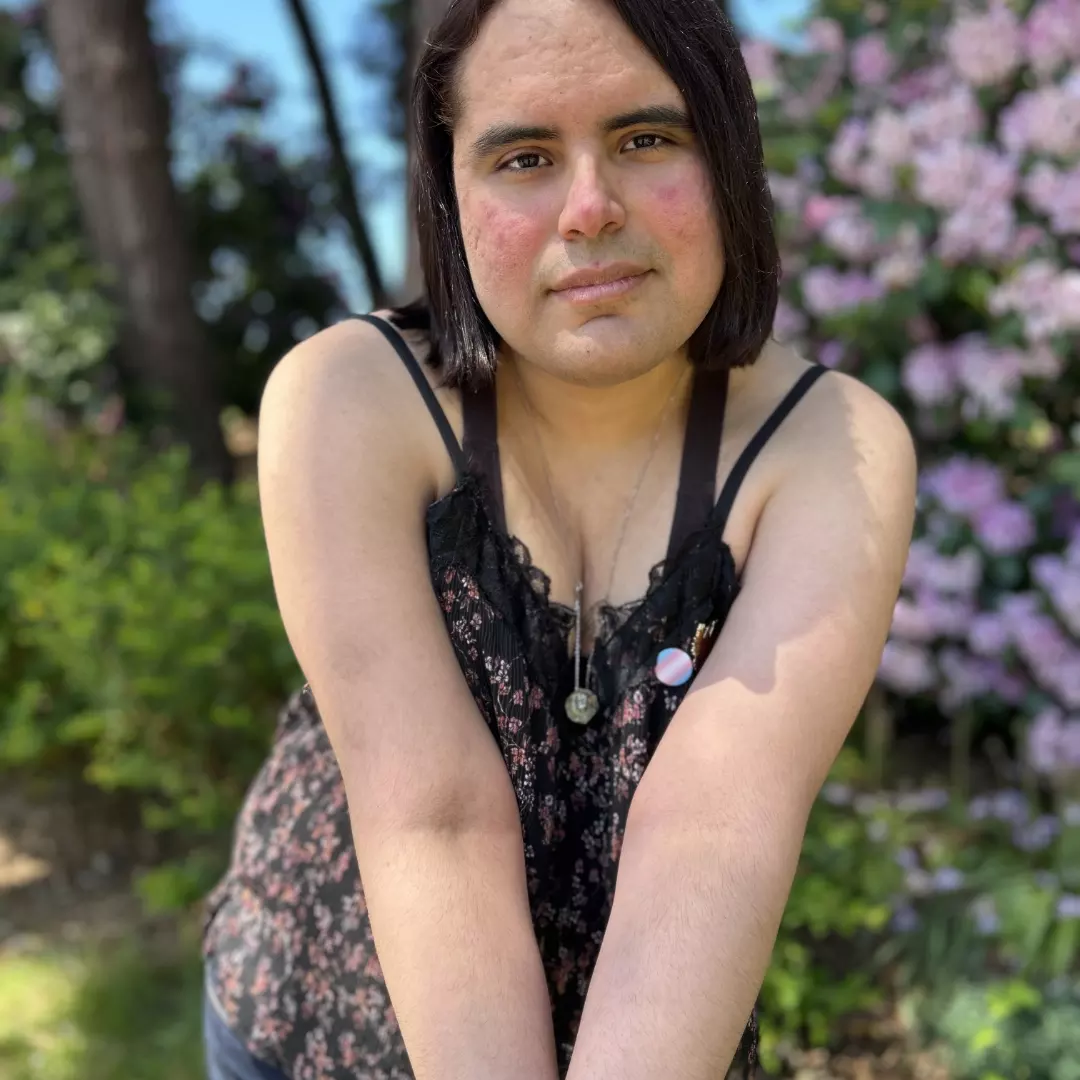 A photo of Angela outdoors leaning forward with arms crossed. She wear a trans pride pin. In the background, sunlight highlights green grass, light pink flowers, and tall trees against a blue sky.