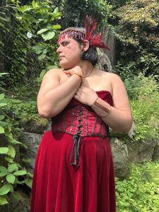 Photo of Angela wearing a red corset and skirt in front of green foliage. Red tasseled beads decorate her forehead and red and black feathers decorate behind her ears.She holds her arms clutched to her chest. Bracelets adorn both her wrists and forearms. Her eyes are closed and she looks to the side.