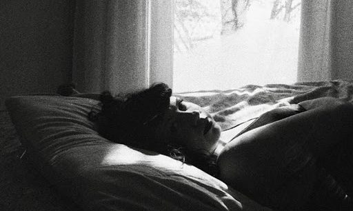 A black and white photograph shows a person laying in a bed by a window. Their face is turned toward us and light from the window shines on their face and pillow behind their head.