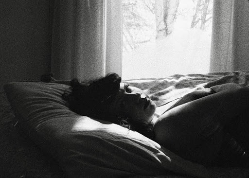 A black and white photograph shows a person laying in a bed by a window. Their face is turned toward us and light from the window shines on their face and pillow behind their head.