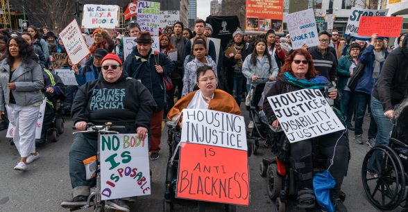 A march of disabled activists in power chairs, scooters, wheelchairs, and walking holding signs. A sweatshirt reads "Fatties Against Fasicsm." Signs read, "No Body is Disposable," "Housing Injustice is Anti Blackness," "Housing Justice is Disability Justice." Disabled activist and ancestor Stacey Park Milbern is in the center of the photo.