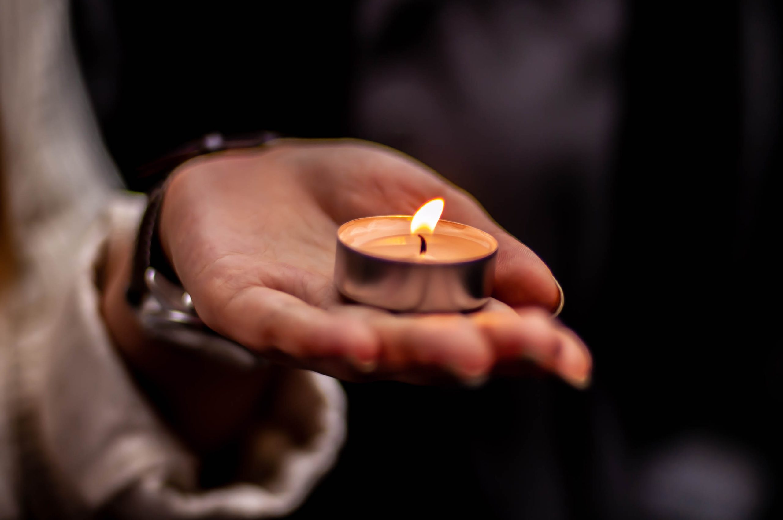 A lit tea light candle in the palm of a person with brown skin and longer oval fingernails.