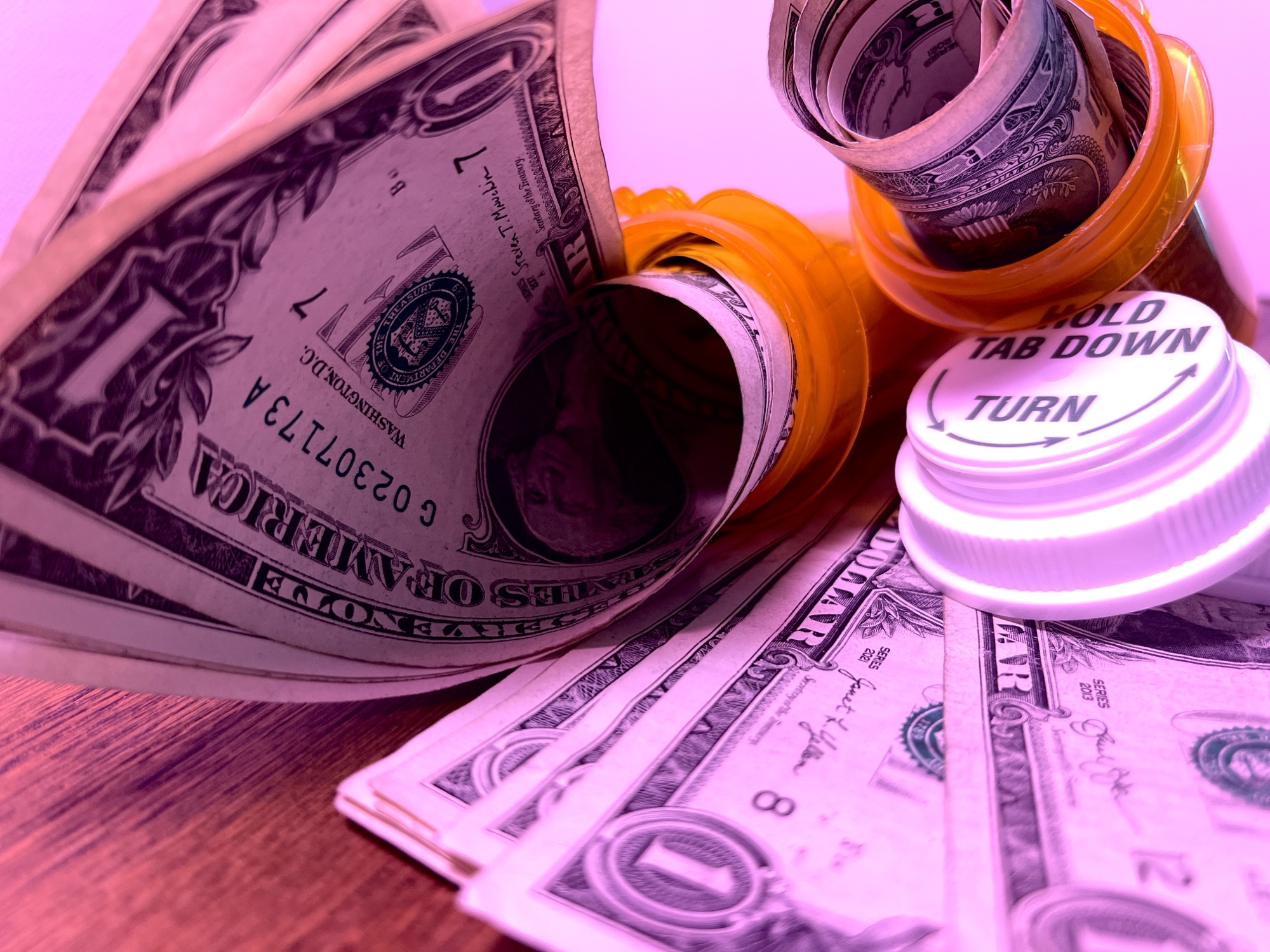 Rolled up dollar bills spiral out of open orange prescription bottles. A lid reads “hold tab down, turn.”