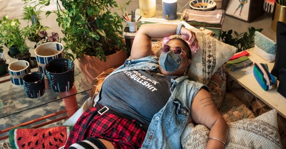 Top-down capture of a non-binary Black person lying back on top of various throw pillows, with one arm arched above their head. They wear tinted purple sunglasses, a KN-95 mask adorned by a double gold chain, and a “Pro-Black Anti-Bullshit” tank top paired with a denim vest and plaid skirt. A pink cane rests next to the person on a watermelon pillow.