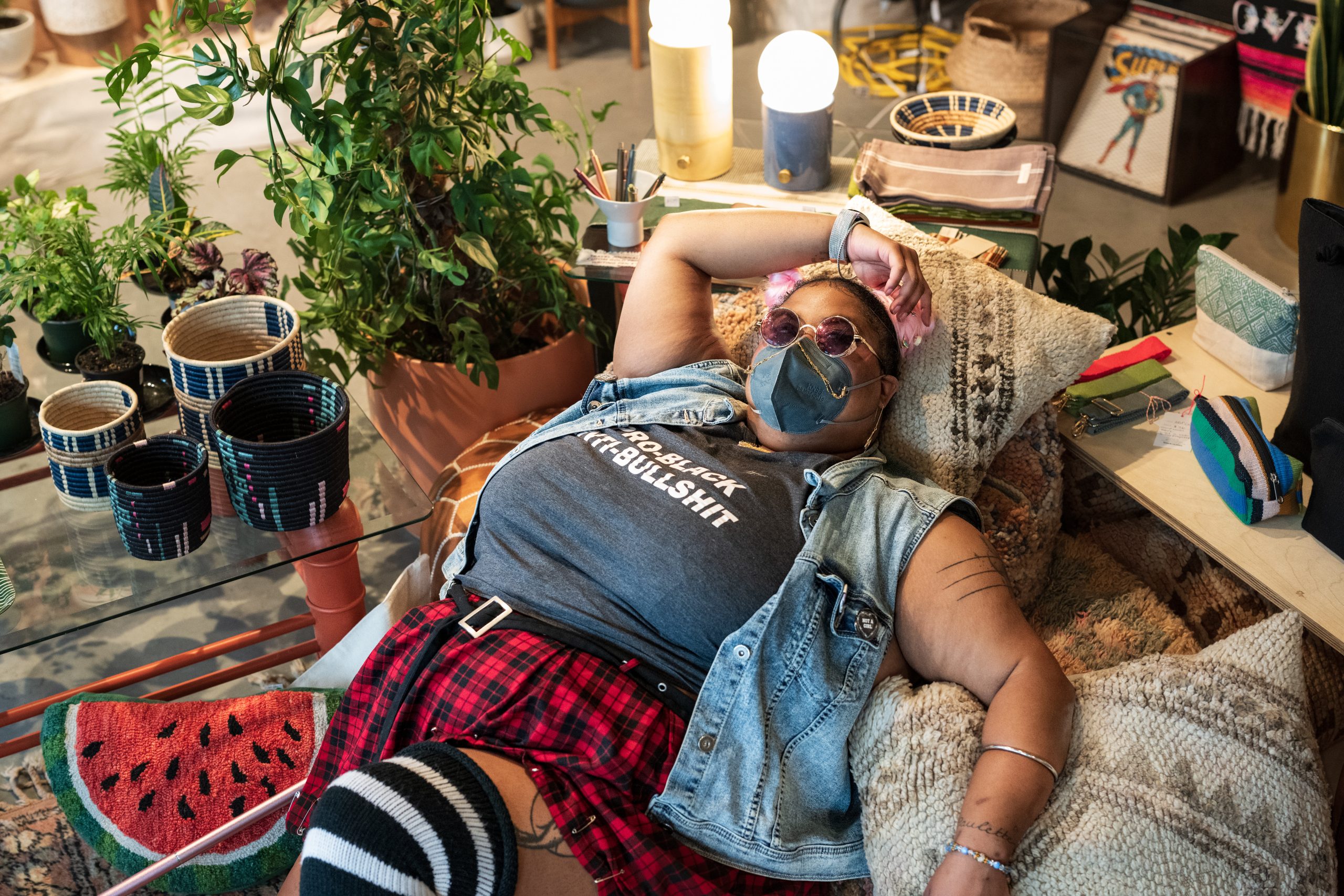 Top-down capture of a non-binary Black person lying back on top of various throw pillows, with one arm arched above their head. They wear tinted purple sunglasses, a KN-95 mask adorned by a double gold chain, and a “Pro-Black Anti-Bullshit” tank top paired with a denim vest and plaid skirt. A pink cane rests next to the person on a watermelon pillow.