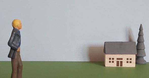 A miniature figure with a hand in their pocket looks over at a miniature house and tree.