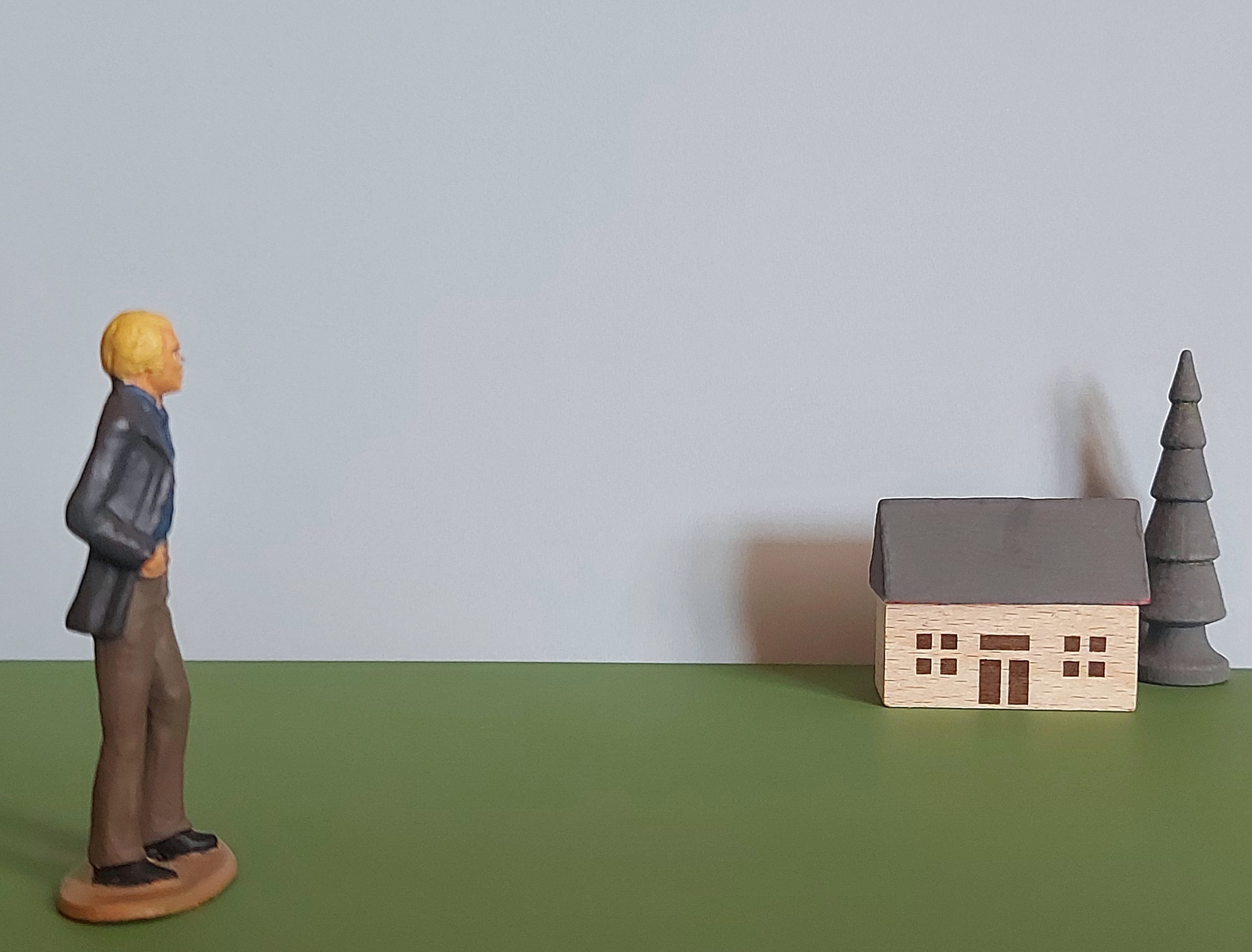 A miniature figure with a hand in their pocket looks over at a miniature house and tree.